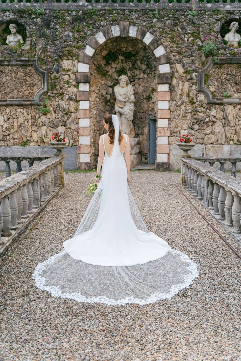 Una sposa in abito bianco con velo, vista di schiena, mentre si trova in un cortile storico con statue di pietra, aiuole fiorite e un muro di pietra con archi.