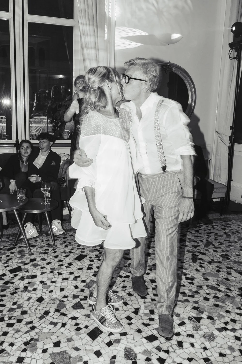 Due persone si baciano durante una festa o un evento sociale, con altre persone sedute sul divano sullo sfondo.