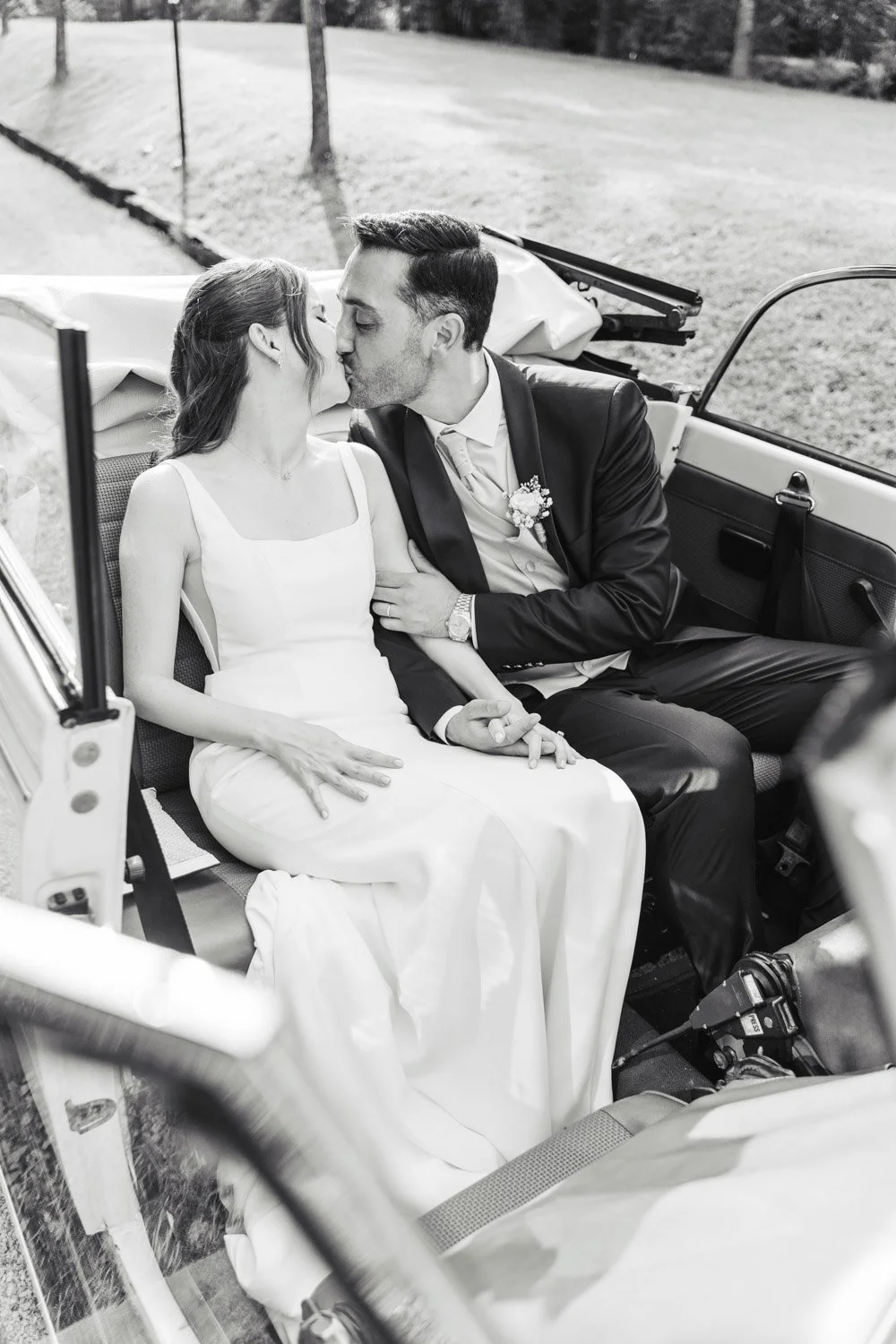 Una coppia di sposi si bacia in un'auto vintage all'aperto.