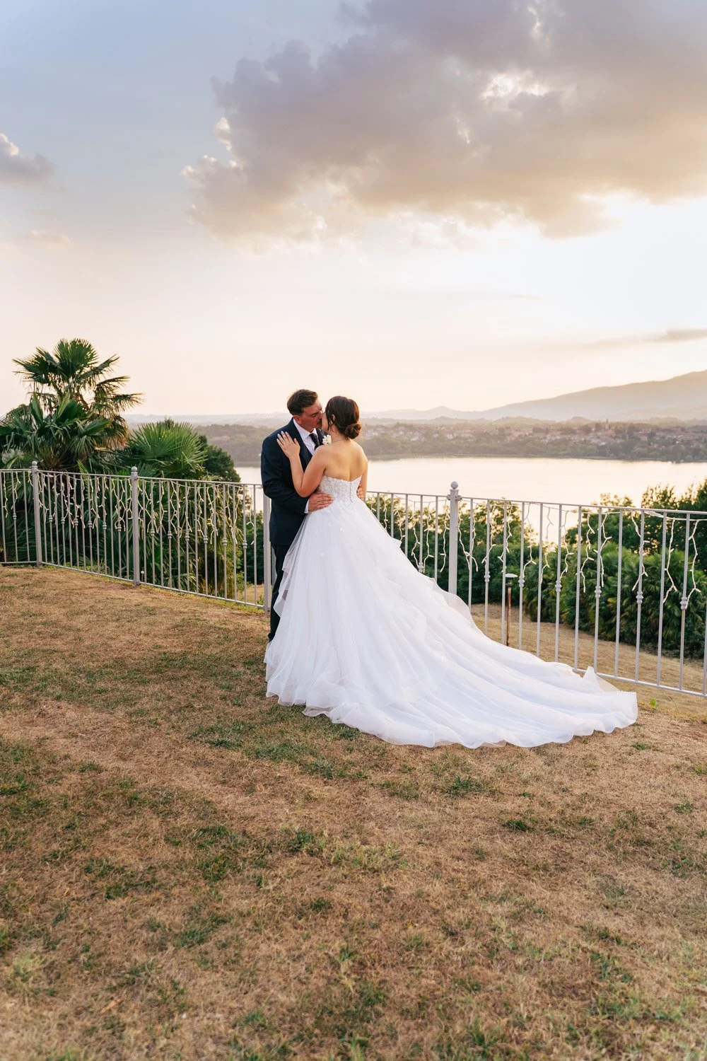 Una coppia di sposi si abbraccia in un matrimonio sulla terrazza durante il tramonto, con uno sfondo di laghi, colline e cielo nuvoloso.