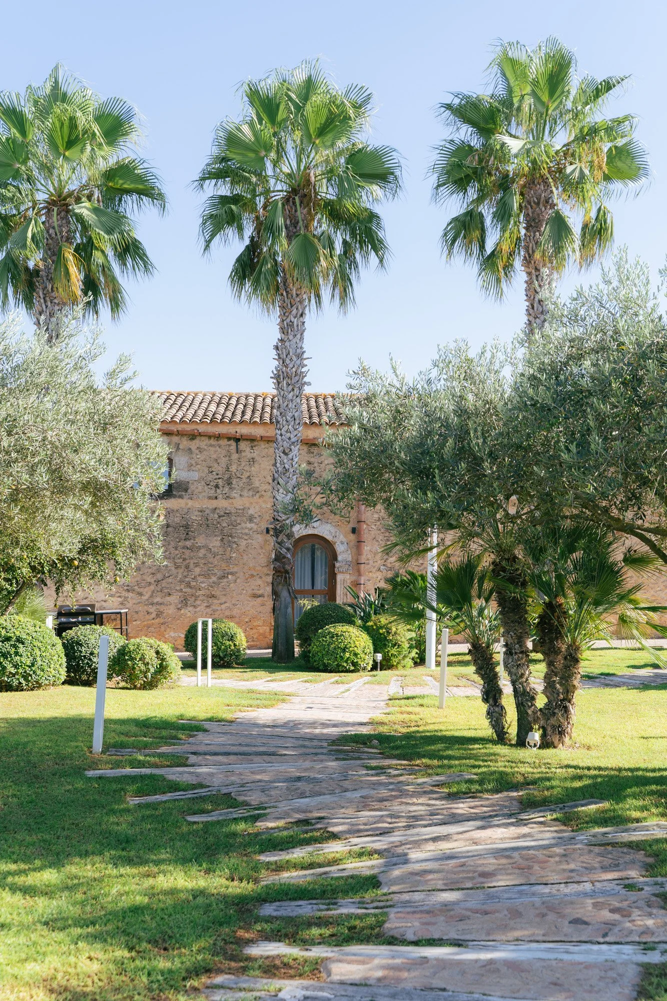 Un cortile con un sentiero di pietra che attraversa un prato verde ben curato, circondato da alberi, tra cui palme e altri arbusti, con un edificio in pietra sullo sfondo e un cielo azzurro.
