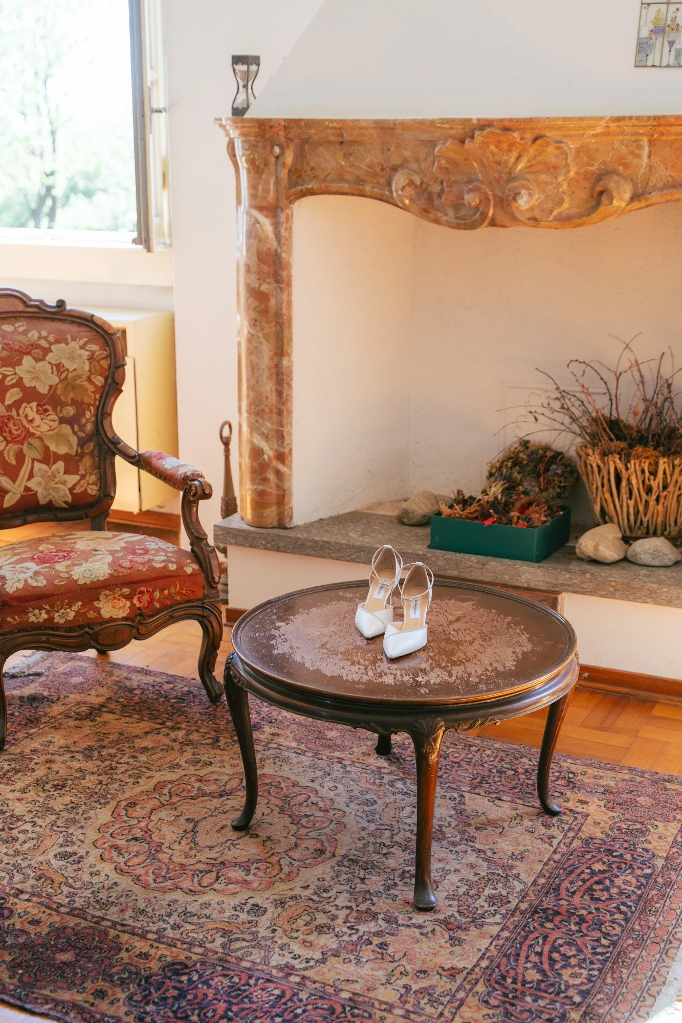 Sala da soggiorno con tappeto, tavolino, sedia imbottita, camino in marmo, vase con fiori secchi e scarpe da sposa