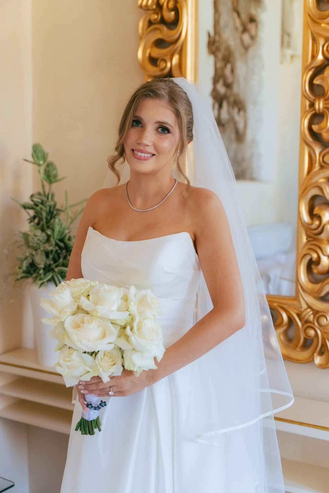 Una sposa sorride e tiene in mano un bouquet di rose bianche, indossa un abito da sposa bianco e una collana, dietro una decorazione dorata e un quadro astratto.