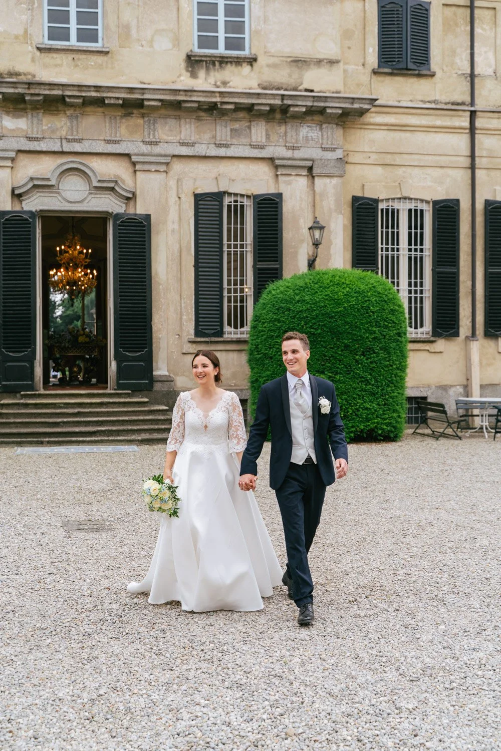 Coppia di sposi in abiti da matrimonio che camminano mano nella mano davanti a una grande casa storica con finestre con persiane nere e un ampio giardino.
