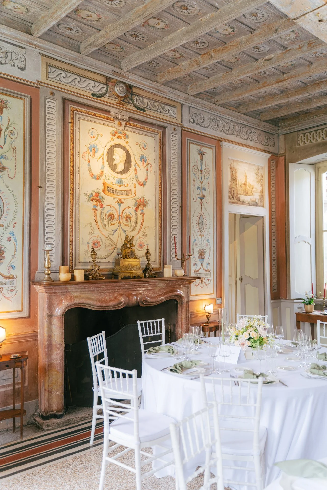 Sala da pranzo elegante decorata con pareti dipinte e un camino in marmo rosso, tavolo imbandito con tovaglia bianca, bicchieri e fiori, e soffitto con travi di legno decorato.