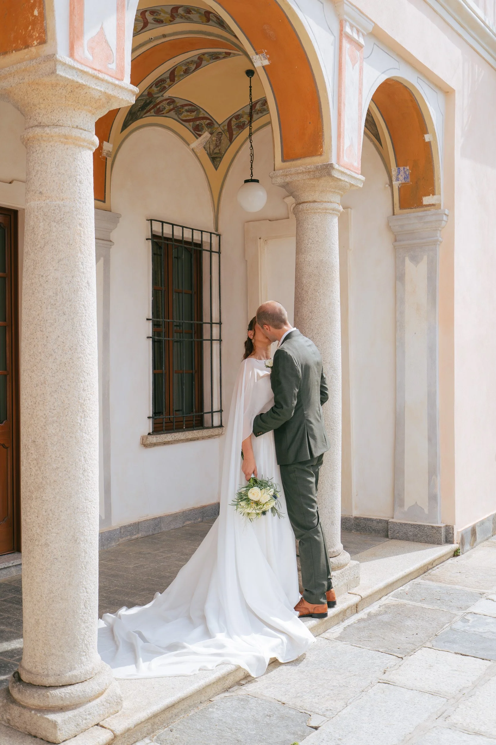 Una coppia di sposi si bacia sotto un portico storico con colonne di pietra e archi dipinti durante un matrimonio.