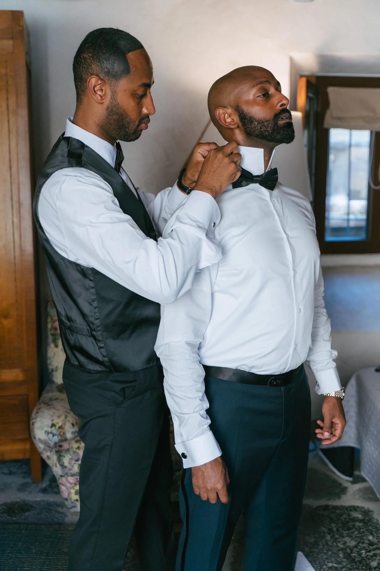 Un uomo sta aiutando un altro uomo ad allacciare una cravatta, entrambi indossano abiti eleganti e si trovano in una stanza con una sedia imbottita e una finestra.