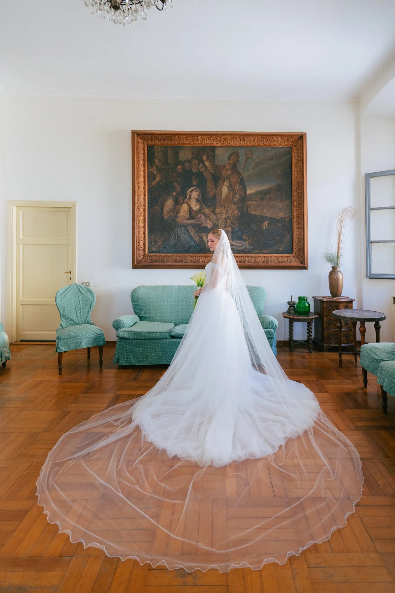 Una donna in abito da sposa bianco con velo, che tiene un bouquet di fiori, si trova in una stanza con pareti bianche, mobili verdi e dipinti. La stanza ha un pavimento di legno e un grande quadro sul muro.
