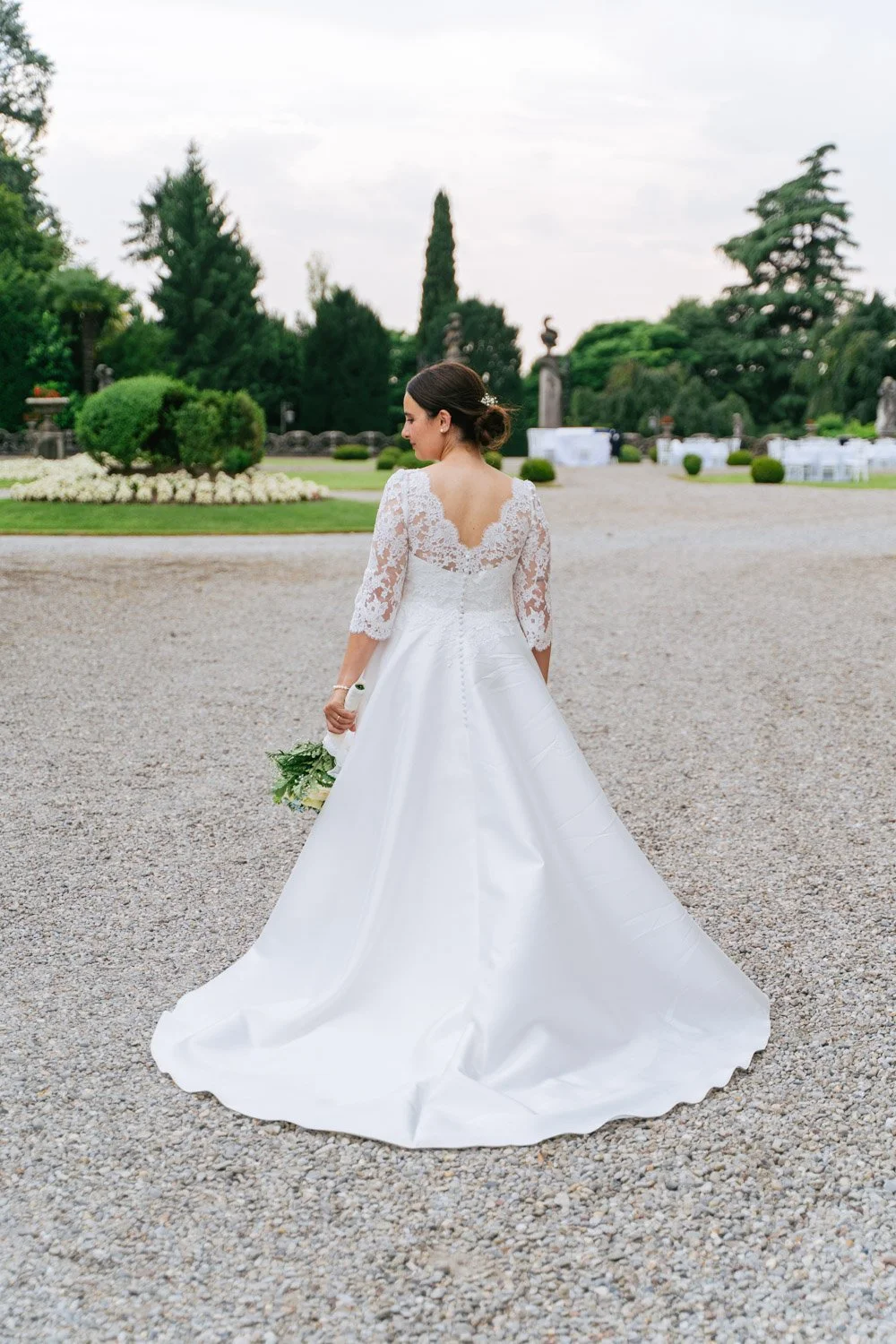 Una sposa in abito da matrimonio bianco con maniche in lace, vista da dietro, che tiene un bouquet verde in una location all'aperto con giardino e decorazioni.