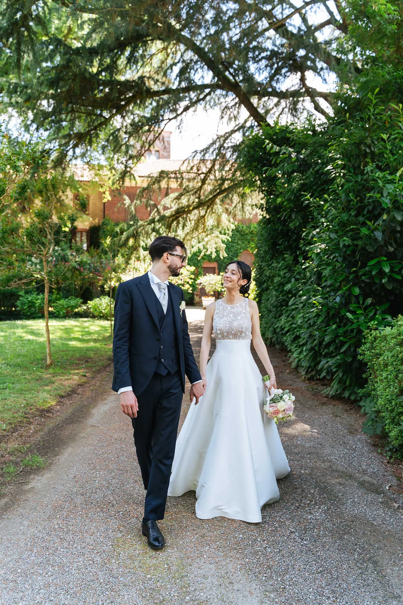 Coppia di sposi che cammina insieme in un giardino, con il manico delle mani, sorridendo e guardandosi, con alberi verdi e un edificio in background.