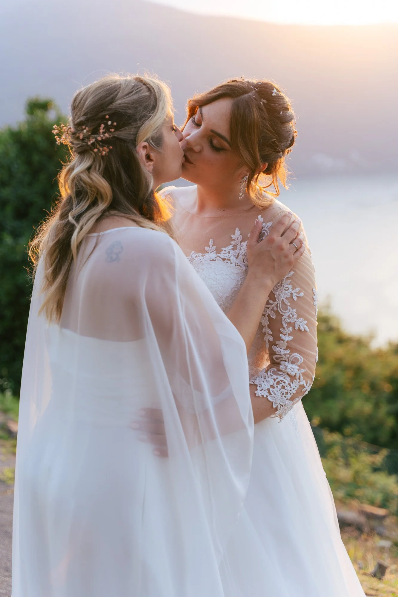 Due donne che si baciano, entrambe in abiti da sposa, con uno sfondo naturale e un cielo al tramonto.