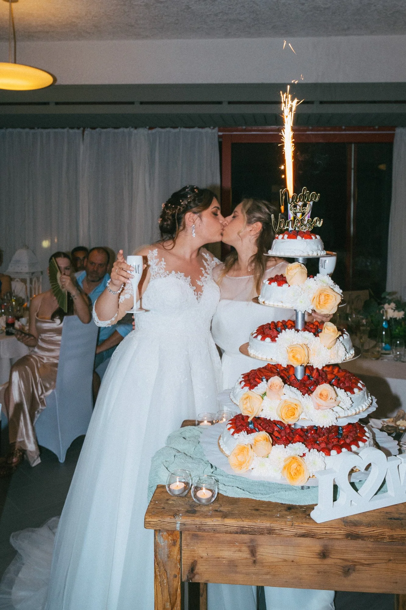 Due donne abbracciate e che si baciano davanti a una torta nuziale con colori vivaci decorata con fiori e una scritta 'Nadia & Vanessa', con un numero di candeline accese in cima, in un ricevimento di matrimonio festeggiato di sera.