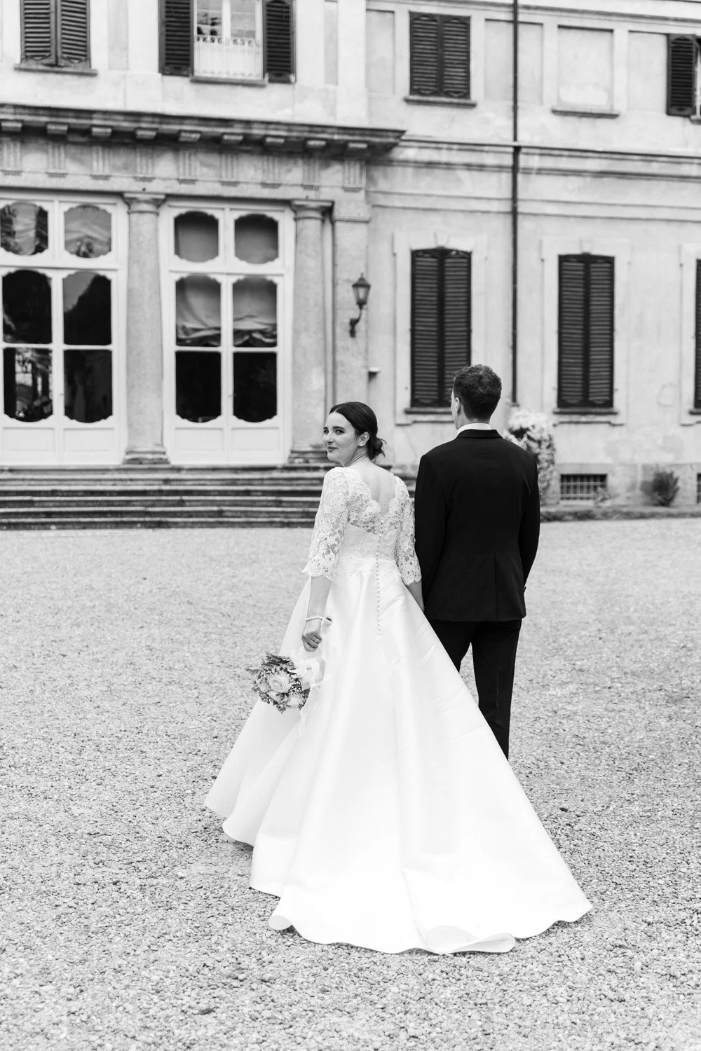 Una coppia di sposi in abiti da matrimonio, la donna tiene un bouquet e guarda indietro, mentre l'uomo si gira di spalle, di fronte a un edificio storico.