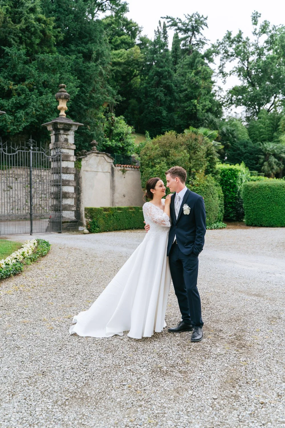 Una coppia di sposi si abbraccia e si sorride vicino a un cancello di ferro e un muro con alberi e cespugli verdi sullo sfondo, in un giardino all'aperto.