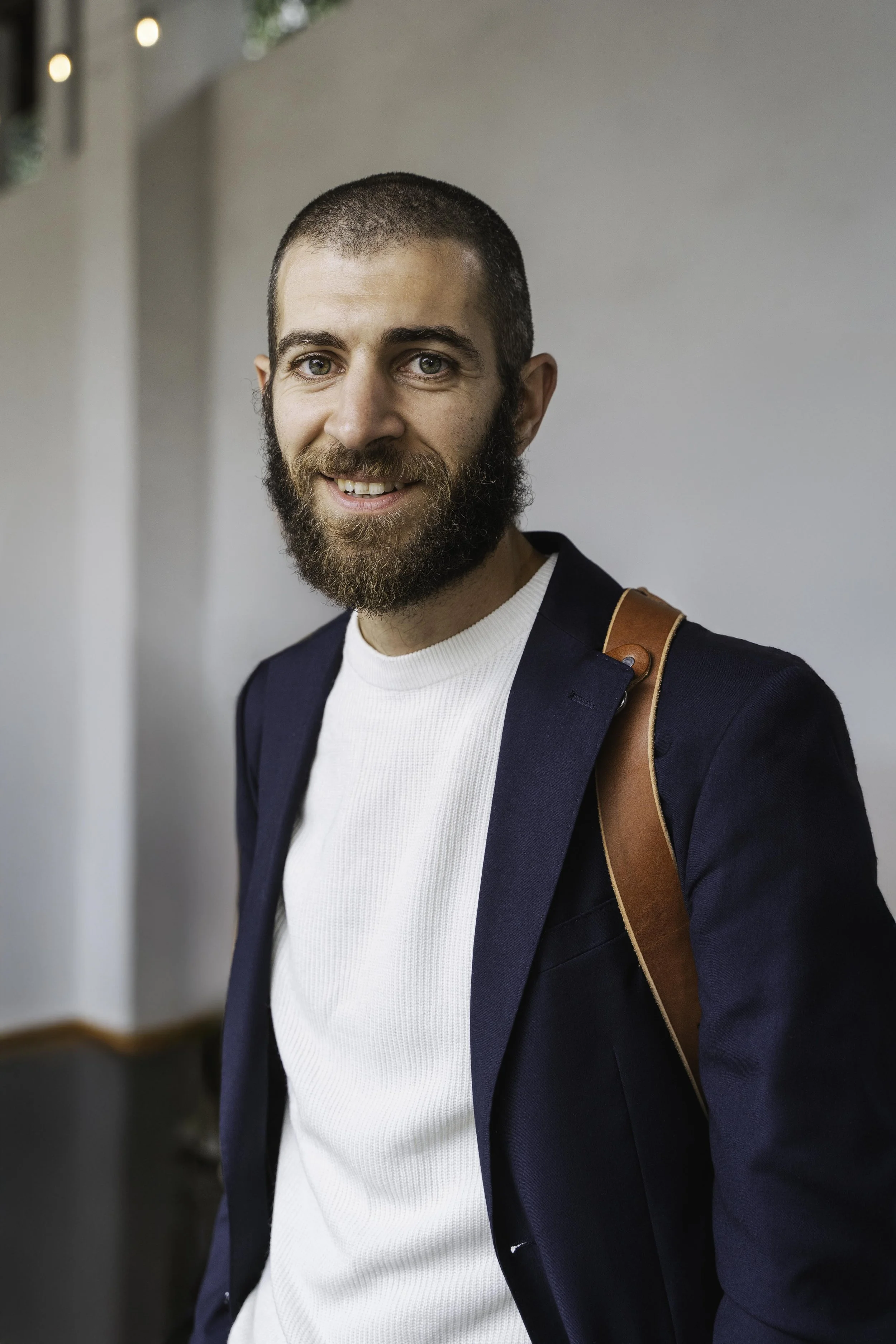Ritratto di un uomo con barba, sorridente, con giacca blu, camicia bianca e tracolla marrone, in un ambiente interno