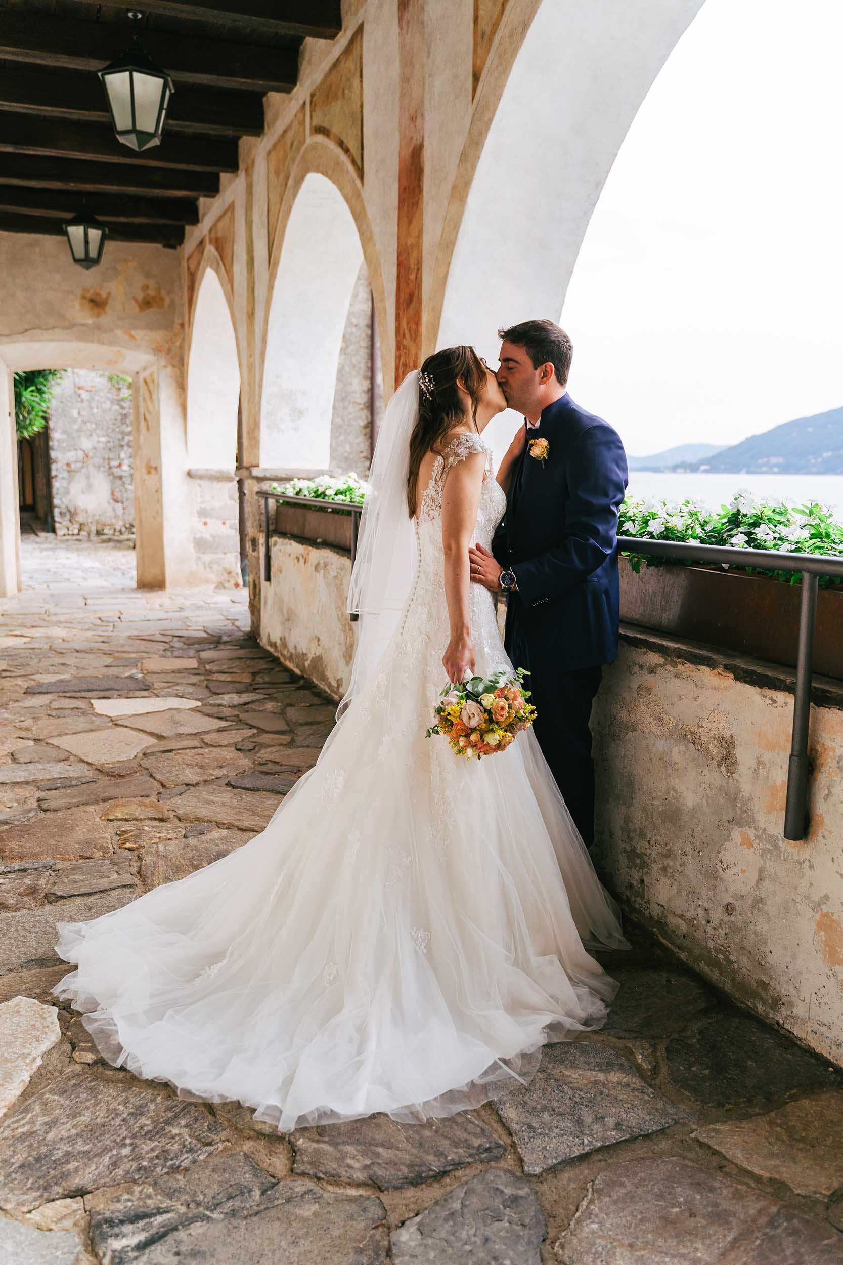 Una coppia di sposi si bacia su un'affacciata in pietra di un edificio storico con archi, con vista su un lago e montagne sullo sfondo. La sposa indossa un vestito da sposa bianco con velo e tiene un bouquet di fiori colorati.