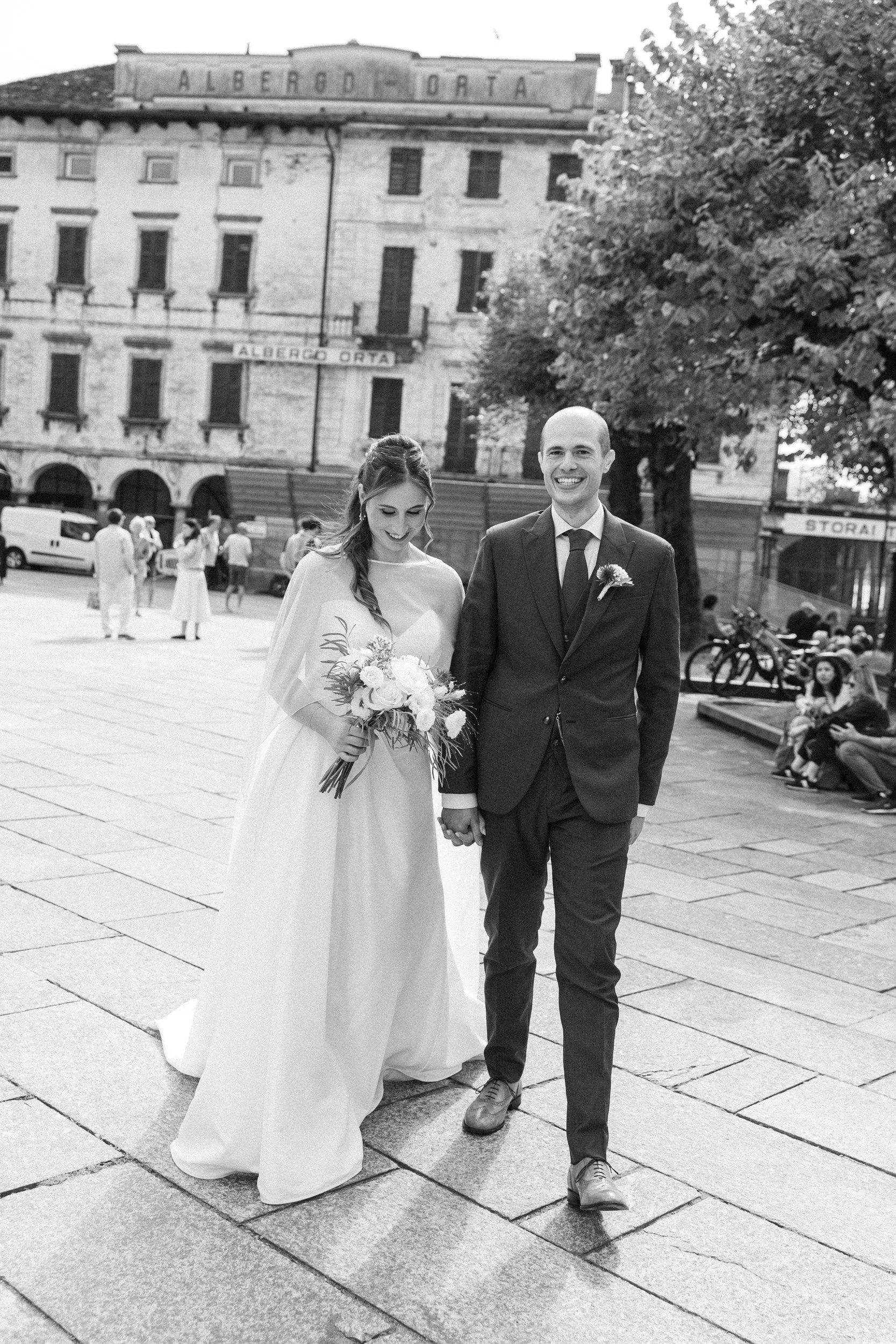 Una coppia di sposi che camminano mano nella mano in una piazza, lei indossa un abito da sposa e lui un completo scuro, entrambi sorridenti.