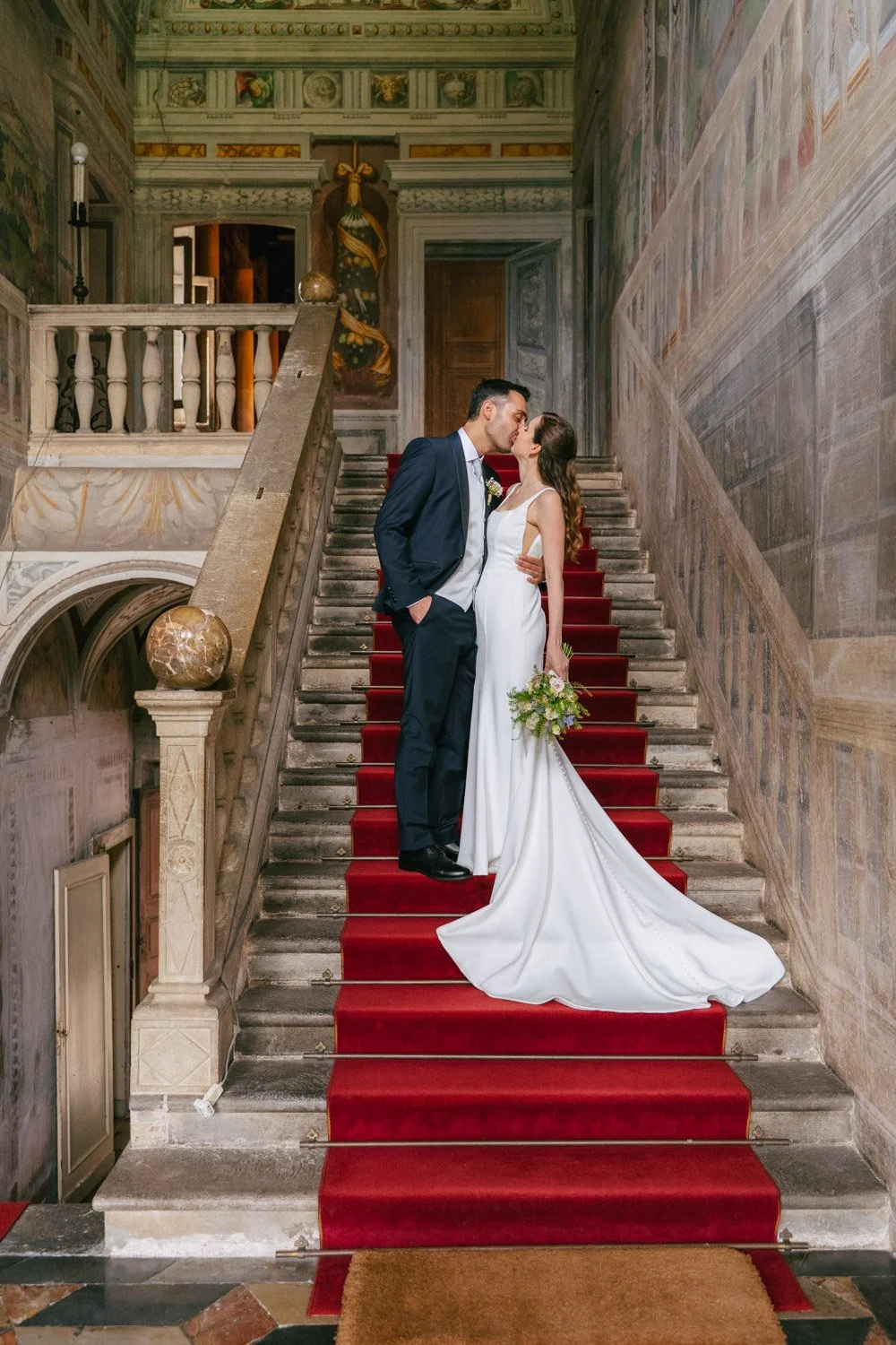 Sposo e sposa che si baciano sulla scala di un palazzo antico, con pareti decorate e un tappeto rosso.