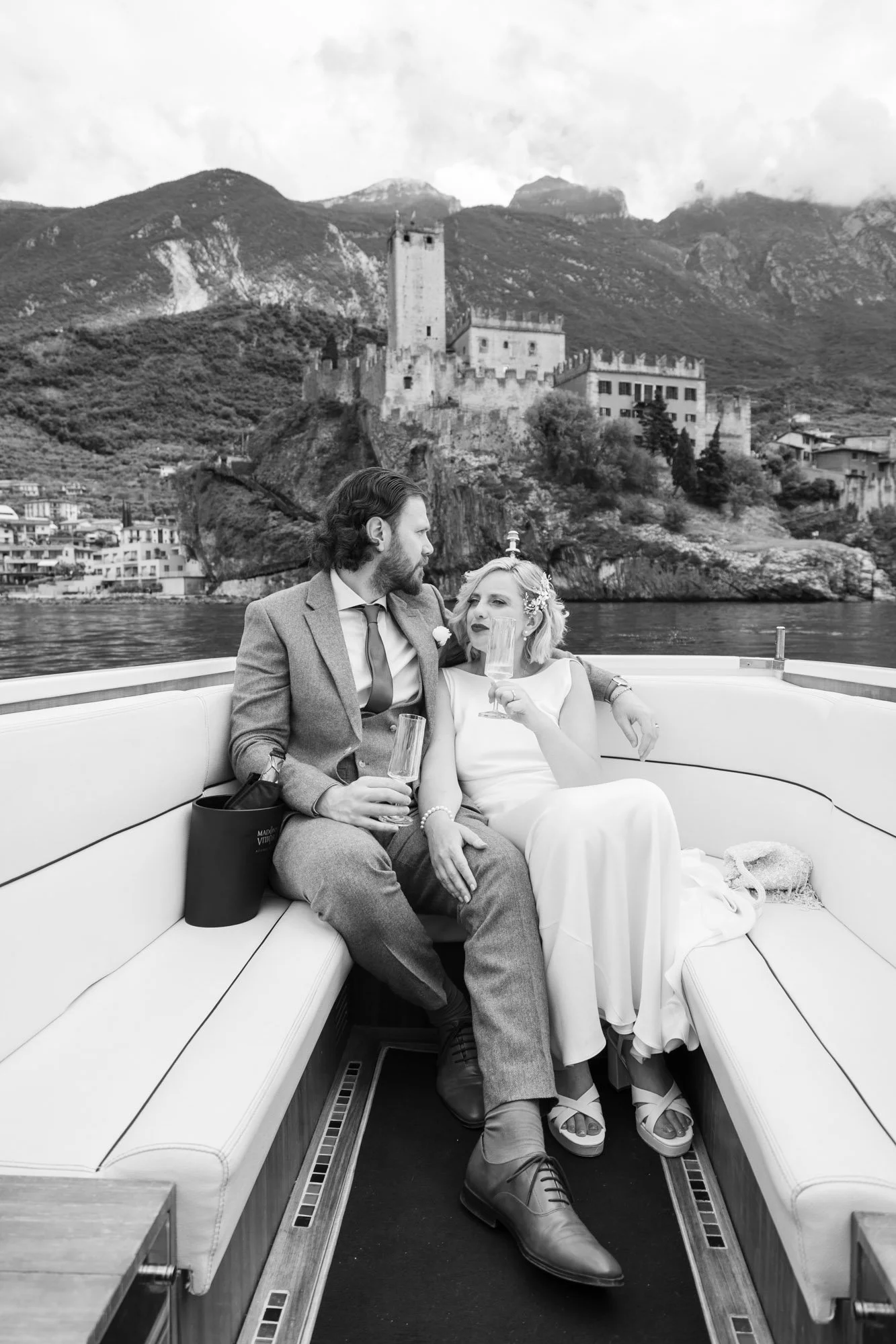 Una coppia di persone sedute su una barca con una vista di un castello medievale e montagne sullo sfondo, in un'immagine in bianco e nero.