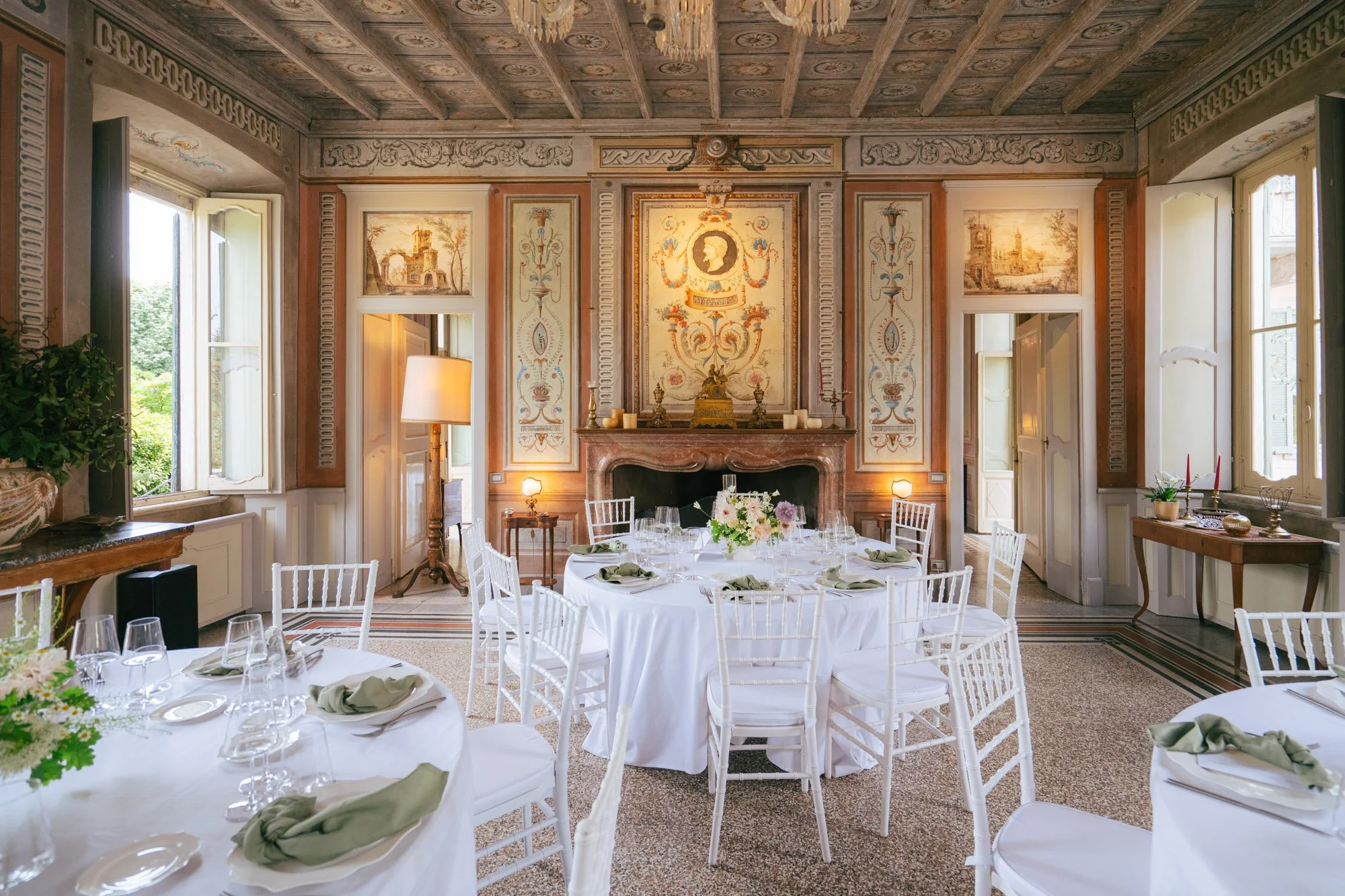 Una sala da pranzo elegantemente arredata con tavoli ricoperti da tovaglie bianche e settati con bicchieri e posate, sedie bianche in stile classico, grandi finestre con tende, affreschi e decorazioni alle pareti, una libreria e un camino in marmo, decorazioni floreali e candelabri.