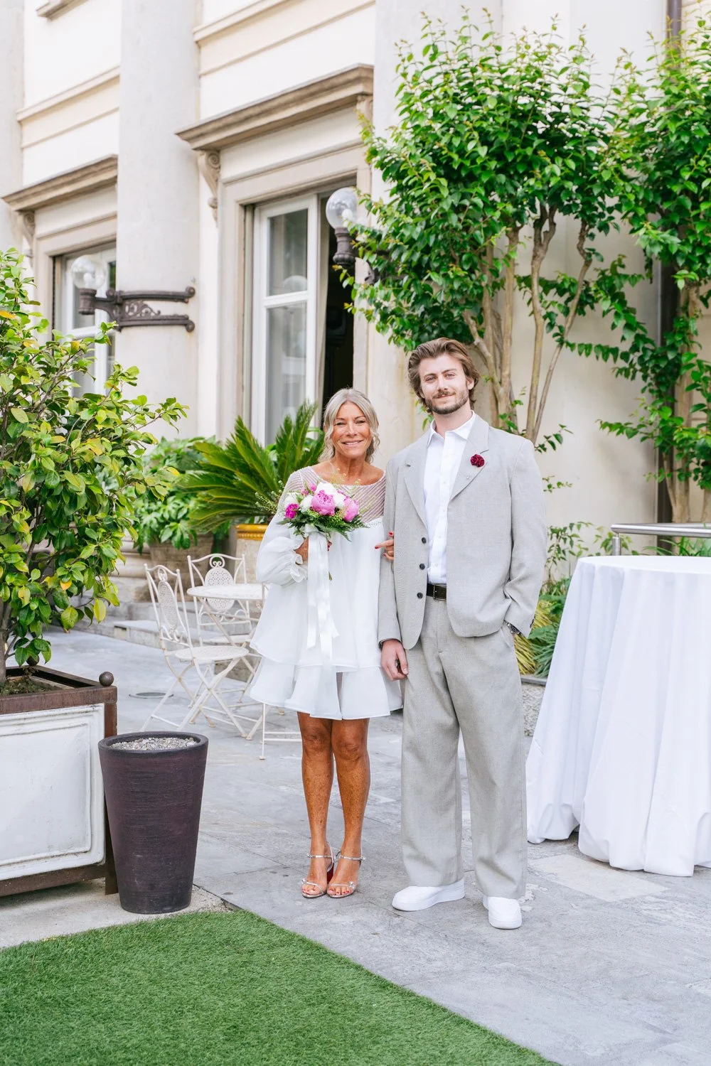 Una coppia, probabilmente sposi, posa sorridente all'aperto in un cortile elegante con piante verdi e mobili in metallo bianco, celebrando un evento speciale.