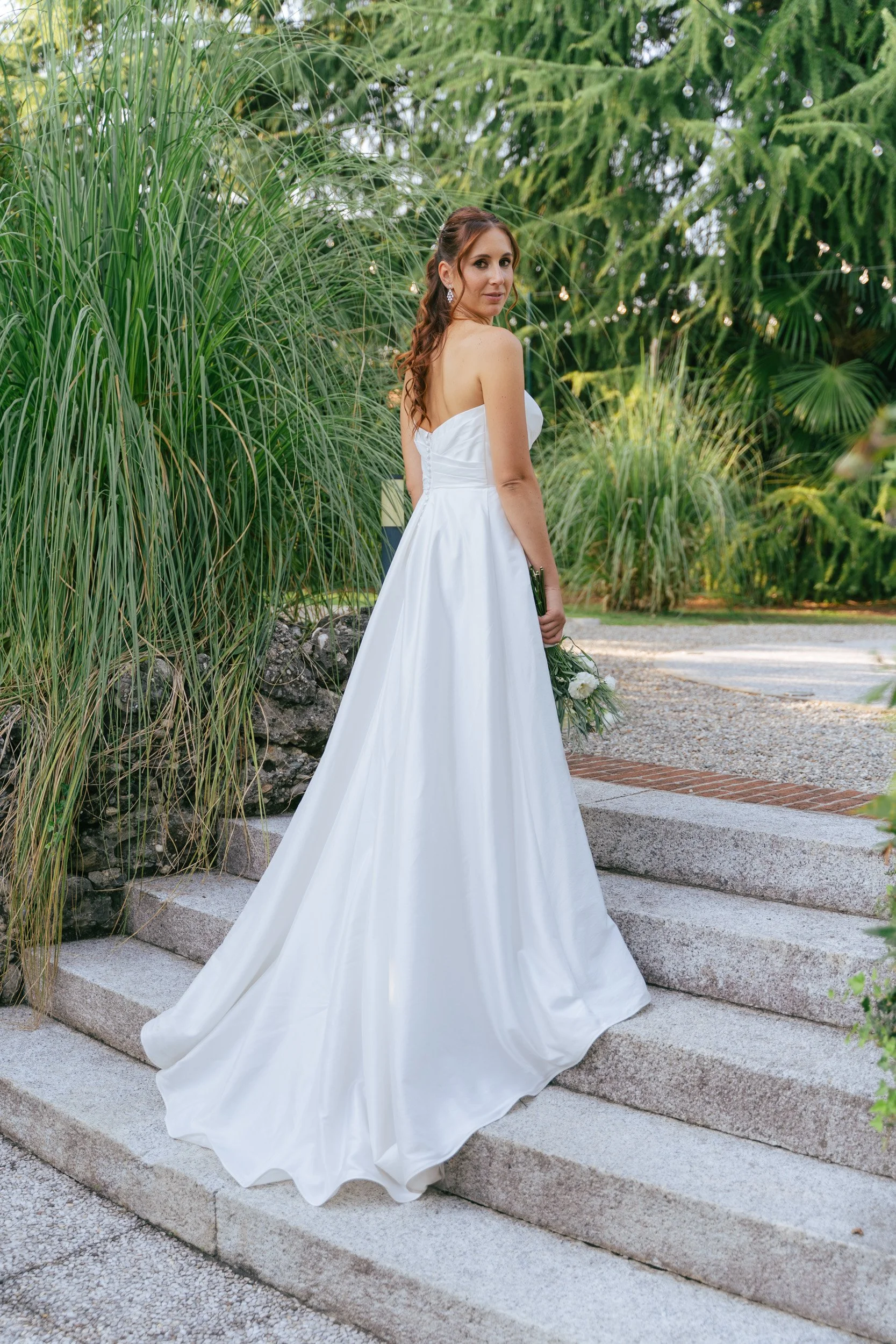 Donna in abito da sposa con sfondo di piante verdi e luci decorative, in piedi sui gradini di pietra.