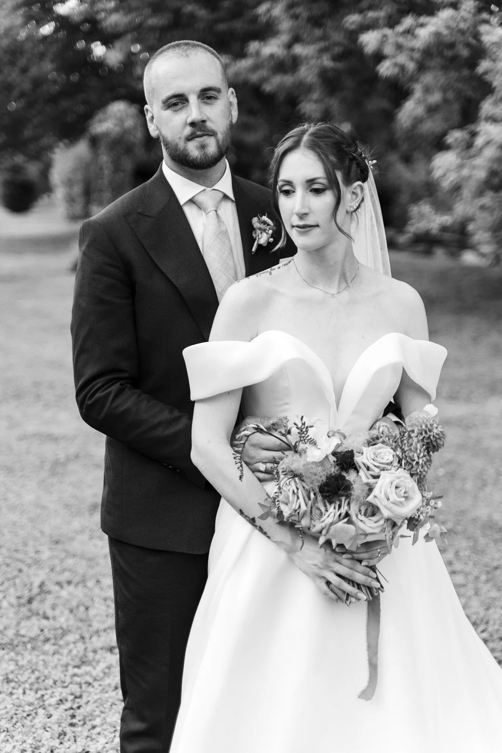 Una coppia di sposi in posa all'aperto, con lo sposo in abito scuro e la sposa in un abito da matrimonio con una corona di fiori, mentre tiene un mazzo di fiori, tutto in bianco e nero.