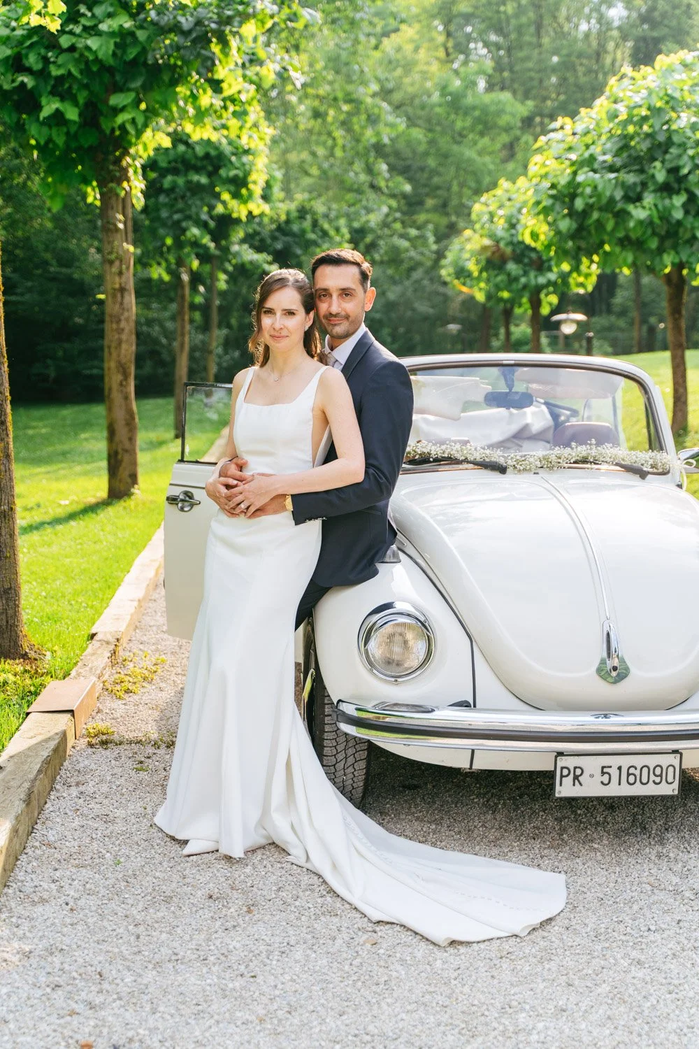 Coppia di sposi in abito da cerimonia davanti a una macchina d'epoca bianca in un parco verde con alberi.