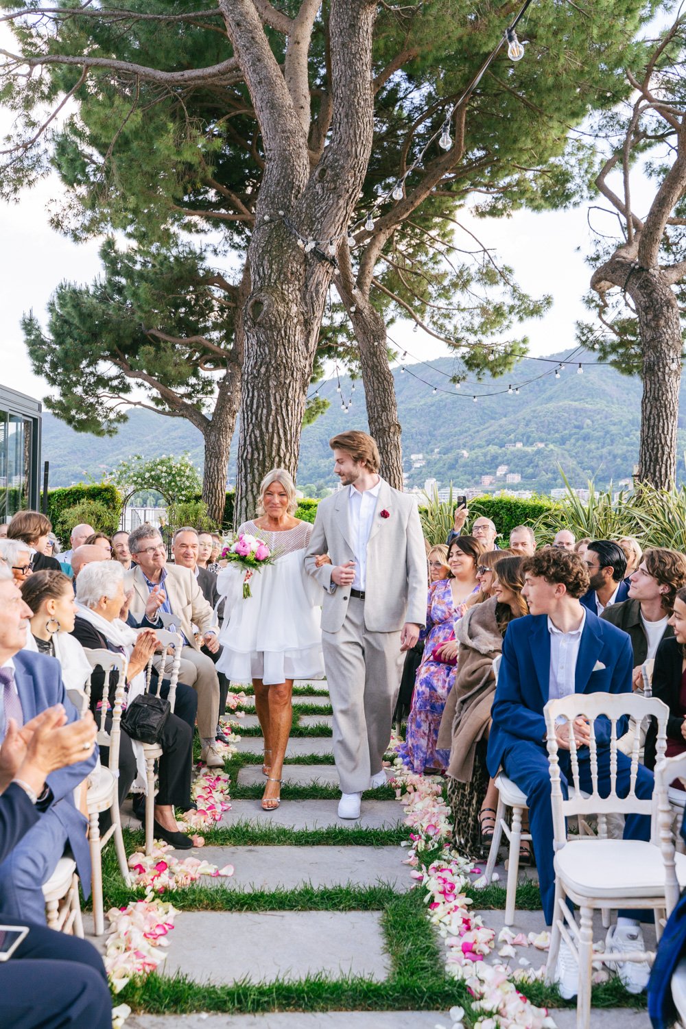 Coppia di sposi che cammina tra gli invitati durante un matrimonio all'aperto, sotto grandi alberi e con decorazioni di luci e petali di rosa.
