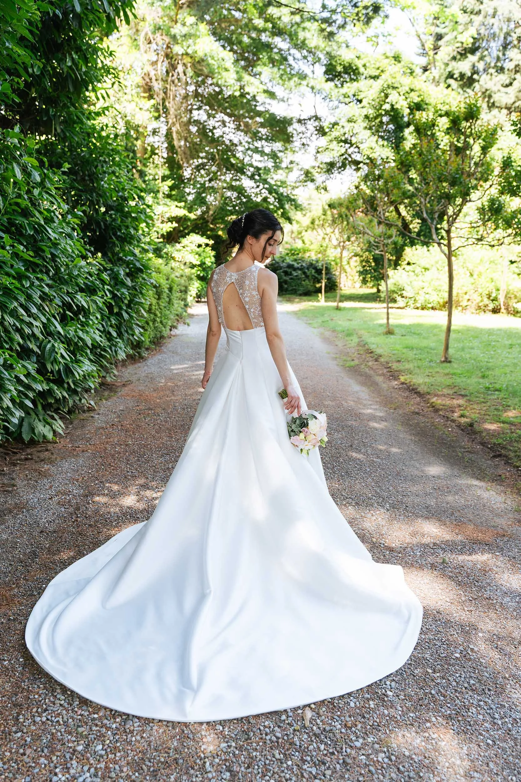 Sposa con abito bianco lungo e schiena scoperta, che tiene un mazzo di fiori, cammina su un sentiero in un parco verde, circondata da alberi e vegetazione.
