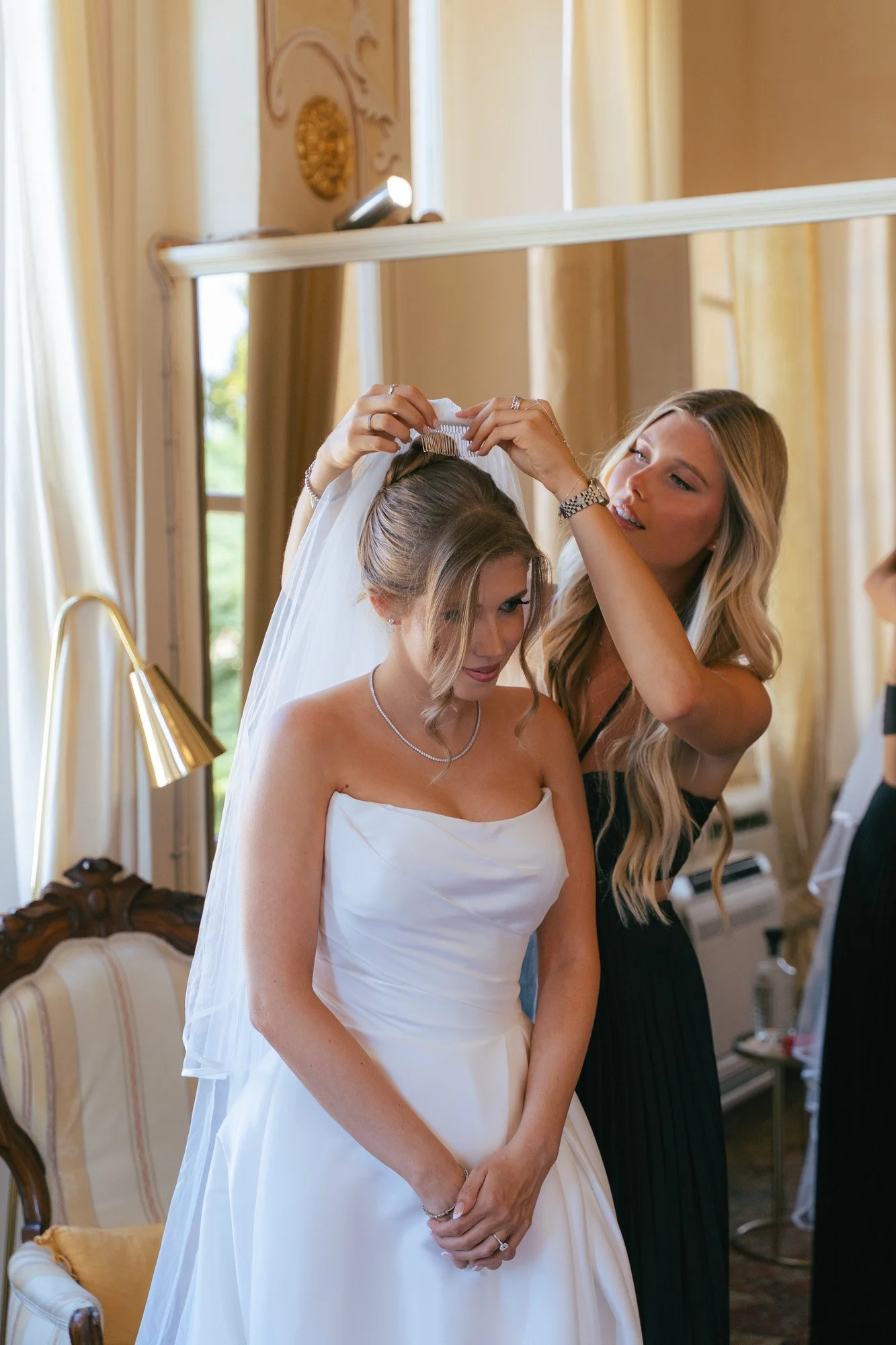 Una donna sta indossando un abito da sposa, mentre un'altra donna la aiuta a mettere il velo in una stanza elegante con tende beige e un grande specchio.