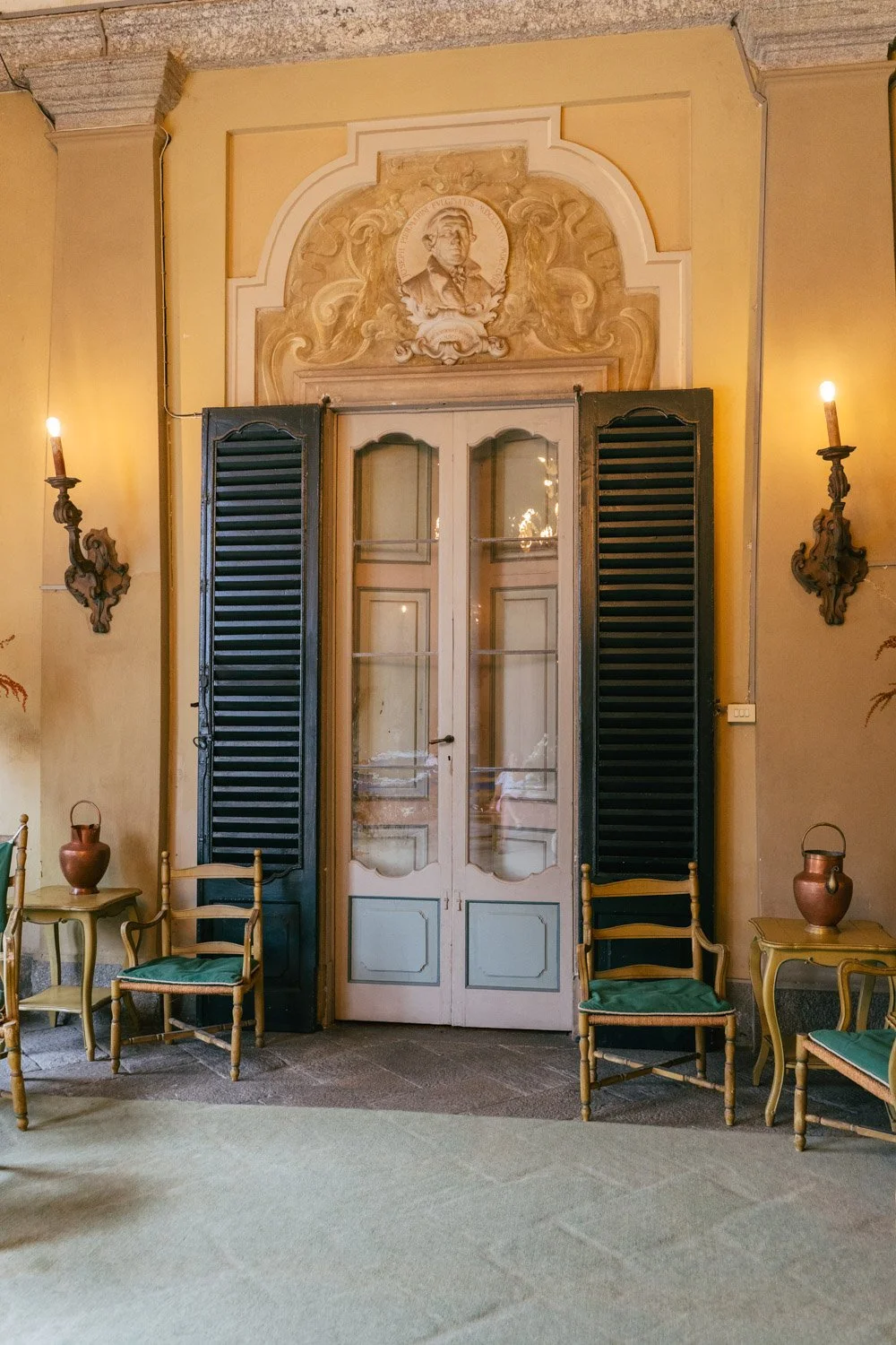 Interno di una stanza elegante con pareti gialle, uno sfondo decorativo con il volto di una persona in rilievo e due lampade da parete ornate. In mezzo, una porta con infissi in vetro e doppie persiane nere, circondata da dettagli architettonici.