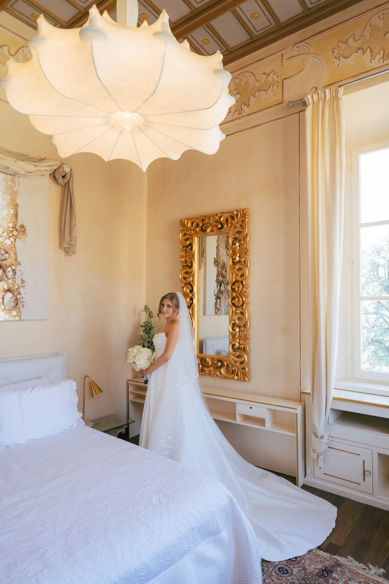 Una sposa in abito bianco tiene un bouquet di fiori chiari in una stanza decorata elegantemente con un grande specchio dorato e tende chiare, illuminata da luce naturale da una finestra.