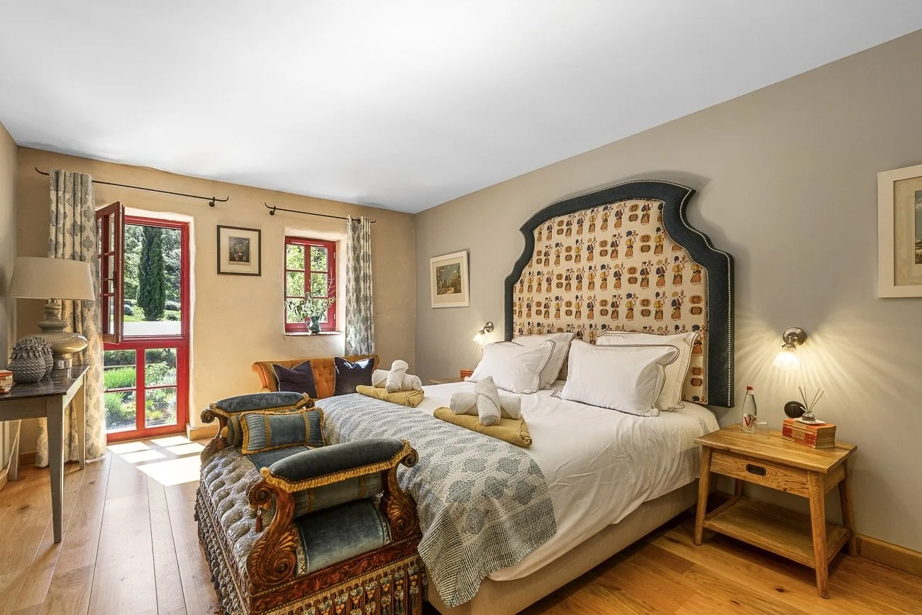 Elegant bedroom with a large bed, ornate headboard, decorative pillows, rolled towels, two bedside tables, a window with red frames, patterned curtains, and wooden floors.