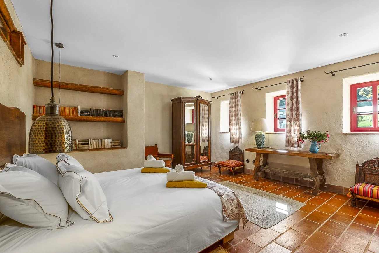 Rustic bedroom with a wooden bed, white pillows and sheets, bedside lamp, wooden shelves with books, a wardrobe with mirrors, red-framed windows, decorative curtains, a wooden table with a lamp and flowers, and terracotta floor tiles.