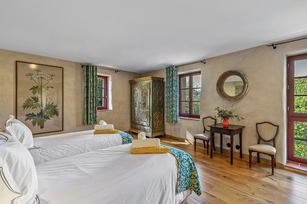 A cozy bedroom with two twin beds adorned with white linens and yellow blankets. An ornate wardrobe stands against the wall. There's a table with a red vase and greenery, two chairs, and large windows with patterned curtains. A round mirror hangs on the wall.
