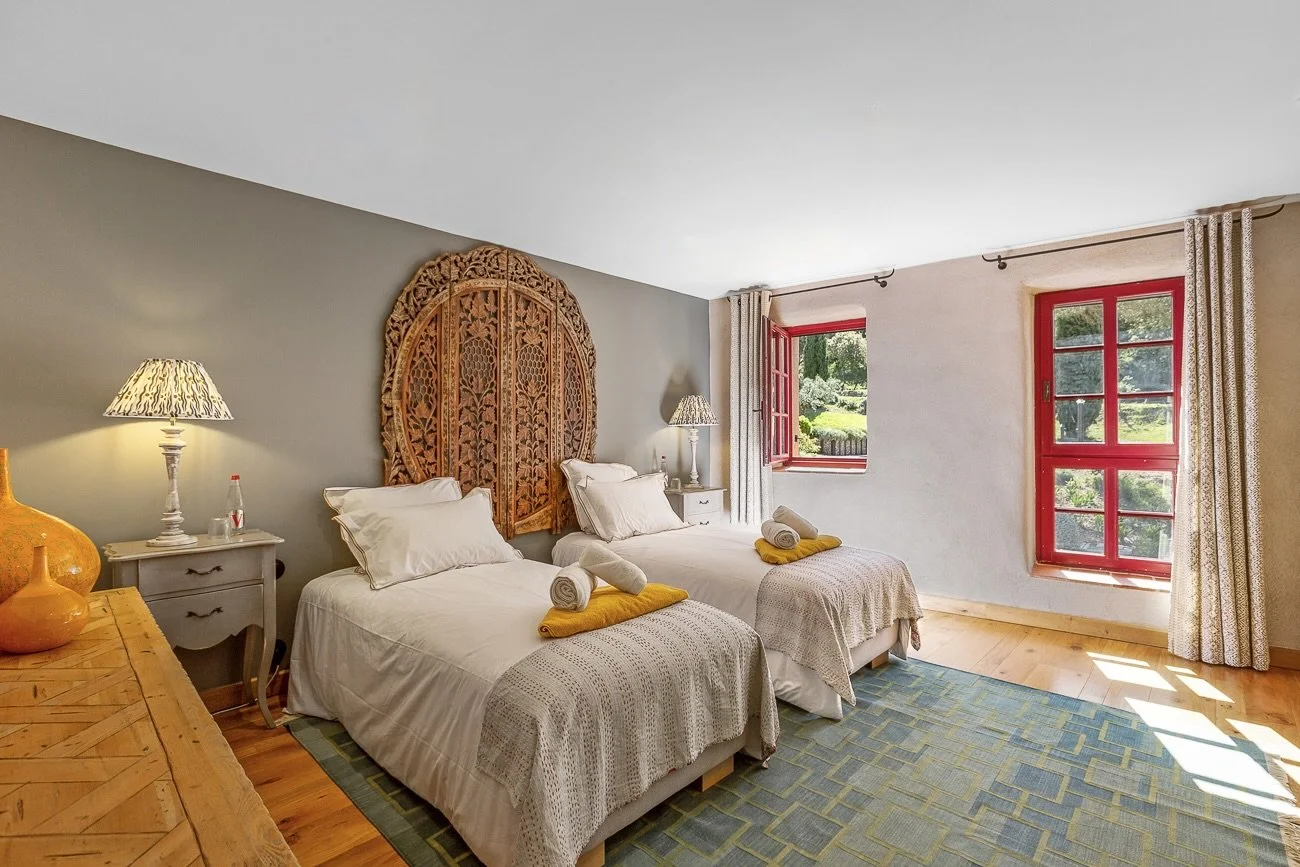 Cozy bedroom with two twin beds, wood-carved headboard, bedside tables with lamps, large window with red frames, and decorative rug.