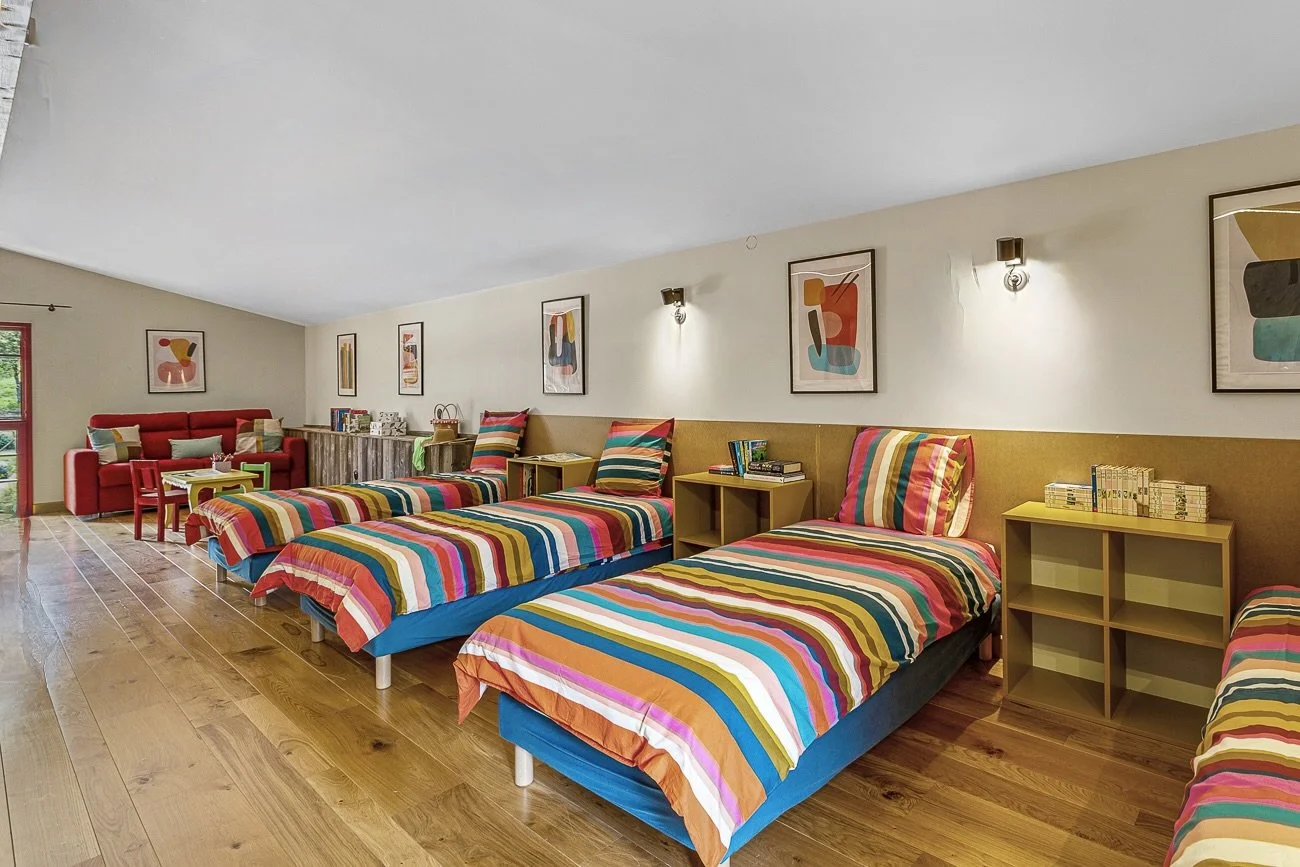 Spacious room with four single beds, featuring colorful striped bedding. The room is adorned with abstract artwork on the walls and has wooden flooring. A red sofa, small tables, and shelves with books and decor items are also present, creating a cozy and vibrant ambiance.