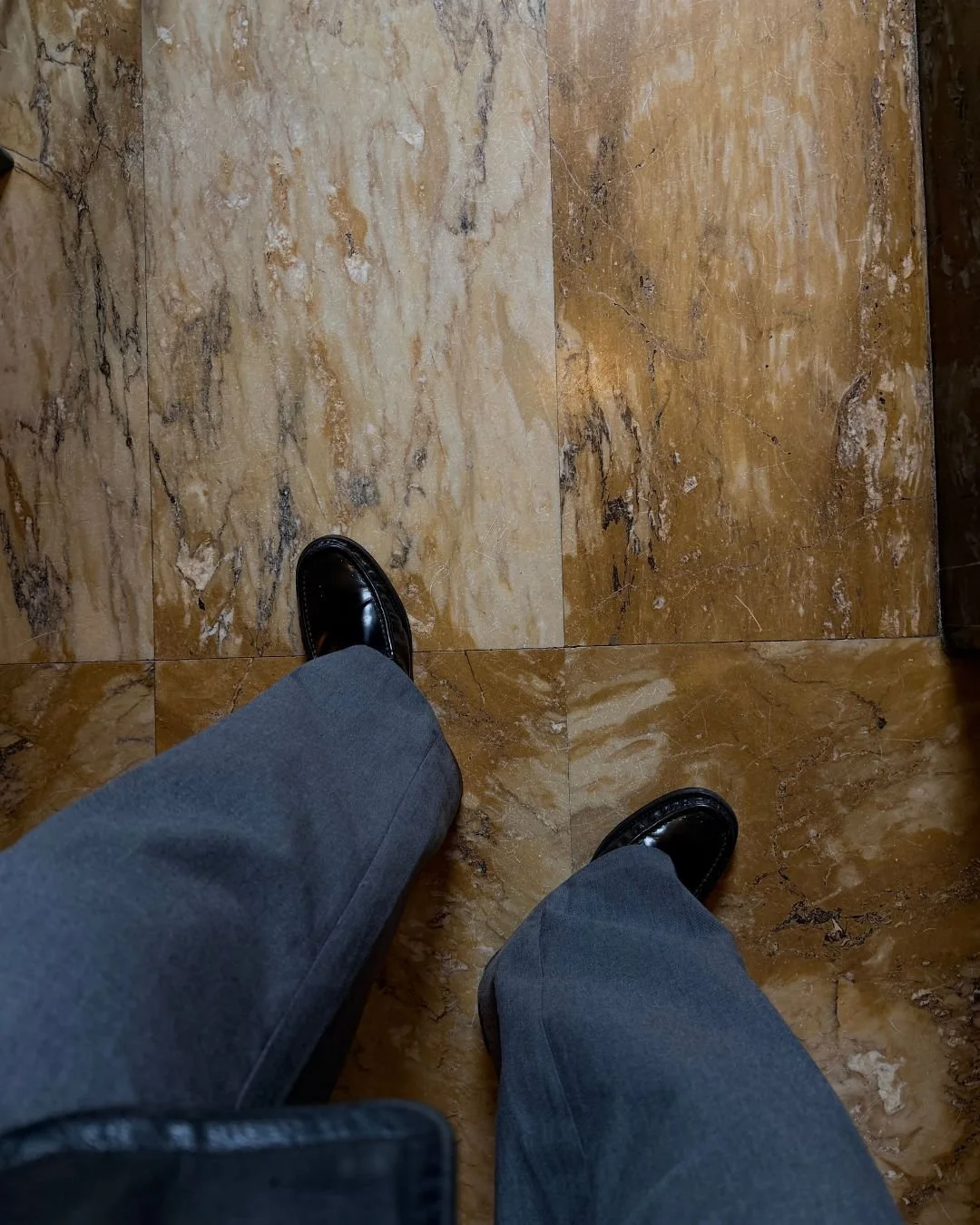Blick nach unten auf den Boden, mit schwarzen Lederschuhen und grauen Hosen, auf einer marmornähnlichen Fliese