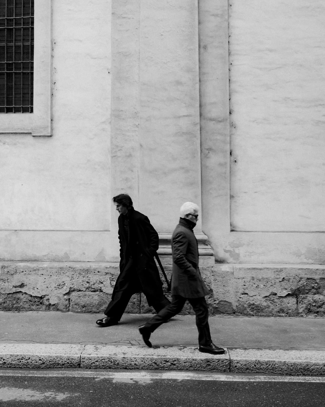 Zwei Männer gehen an einer weißen Wand entlang, einer trägt einen schwarzen Mantel und der andere einen grauen Mantel mit Gesichtsschutzmaske.