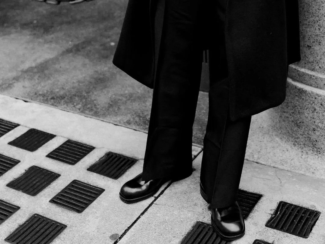 Der untere Körper eines Menschen in dunkler Kleidung, schwarzen Schuhen und einer schwarzen Jacke, der auf einer Bürgersteig mit Gitterabläufen steht.