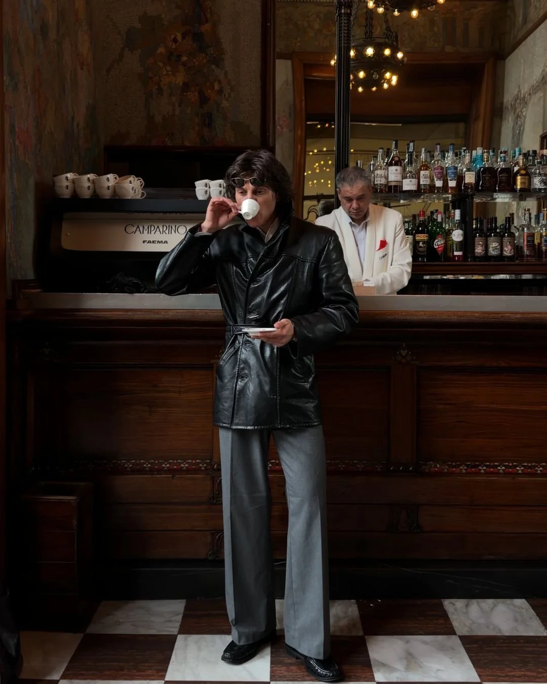 Eine Person mit schwarzem Ledermantel trinkt Kaffee in einem Café, im Hintergrund eine Bar mit Spirituosen. Es ist dunkel und gemütlich eingerichtet.