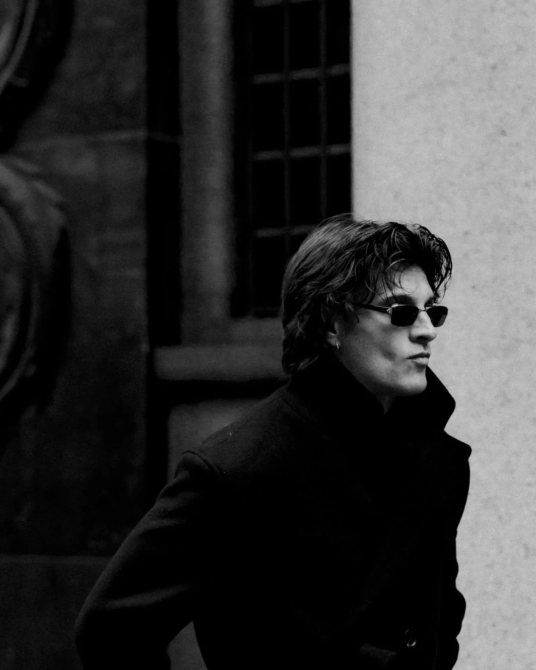 Schwarz-weiß-Foto eines Mannes mit mittellangem Haar, Sonnenbrille und dunklem Mantel, der an einer Wand entlanggeht.