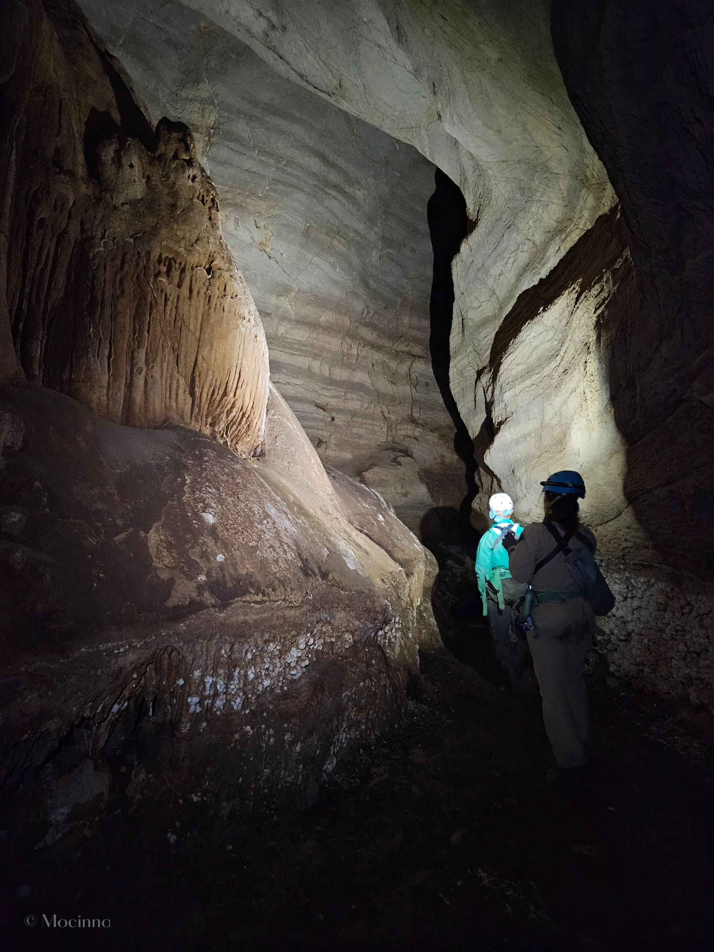 People exploring a large cave with high ceilings and rock formations, wearing helmets and outdoor gear.