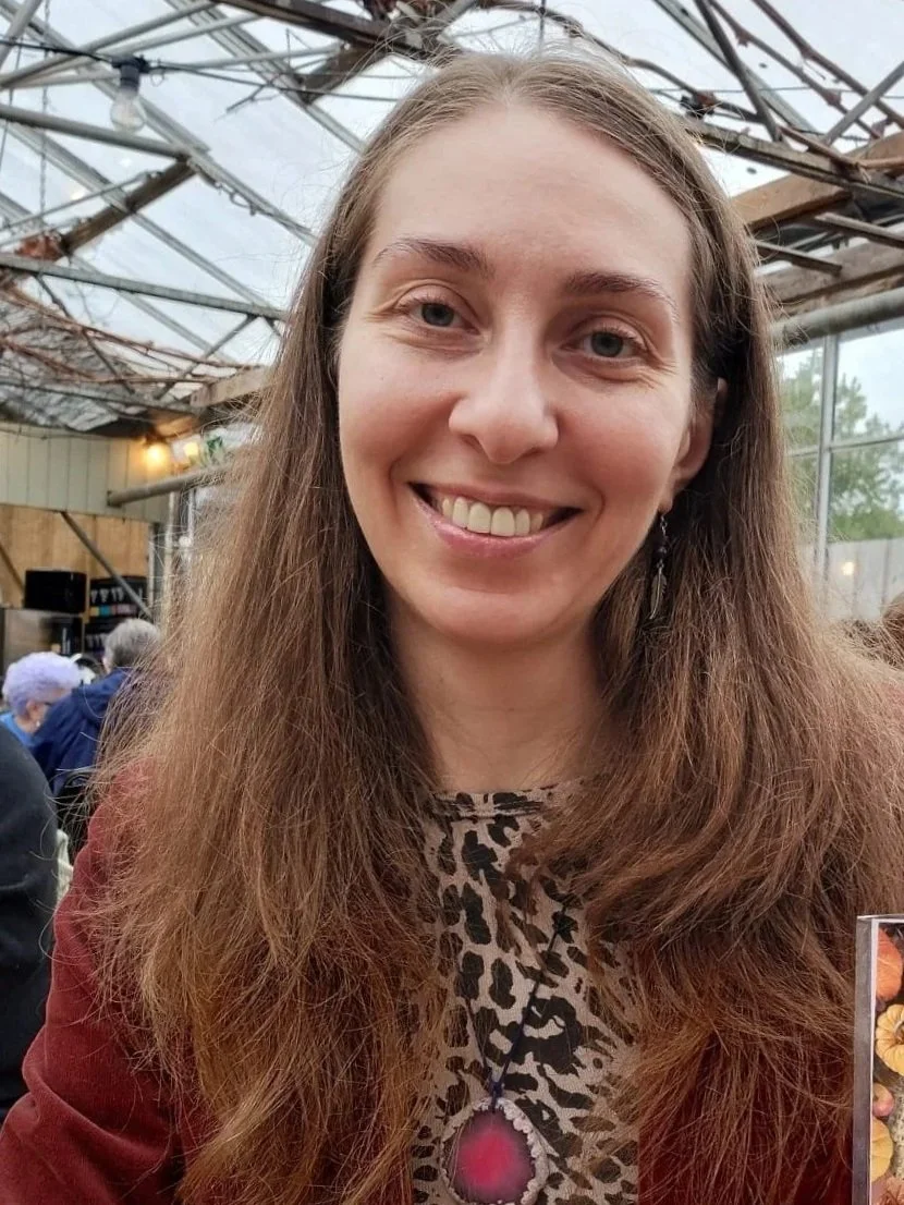 A woman with long brown hair, smiling, wearing a leopard print top, a necklace with a large pink pendant, and an earring. She is indoors, possibly in a greenhouse or similar setting with a metal and glass roof.