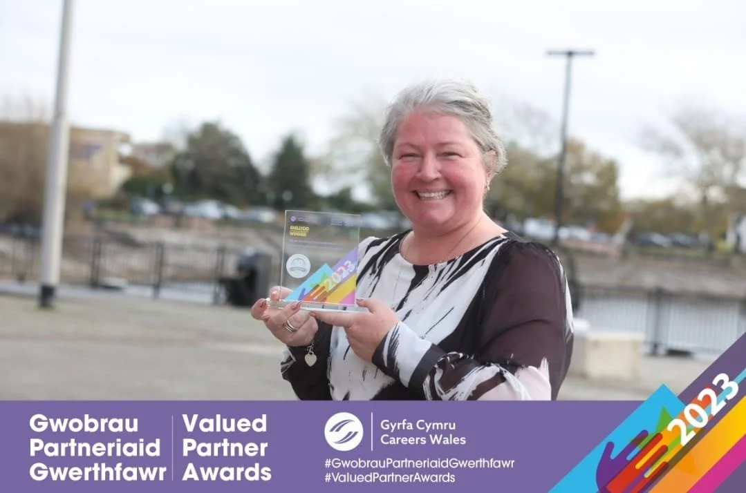 A smiling woman holding an award plaque outdoors, with a waterfront and trees in the background, and an overlay banner that reads 'Gowbrau Partneriaid Gwerthfawr Valued Partner Awards 2023' and 'Gyfry Cymru Careers Wales'.