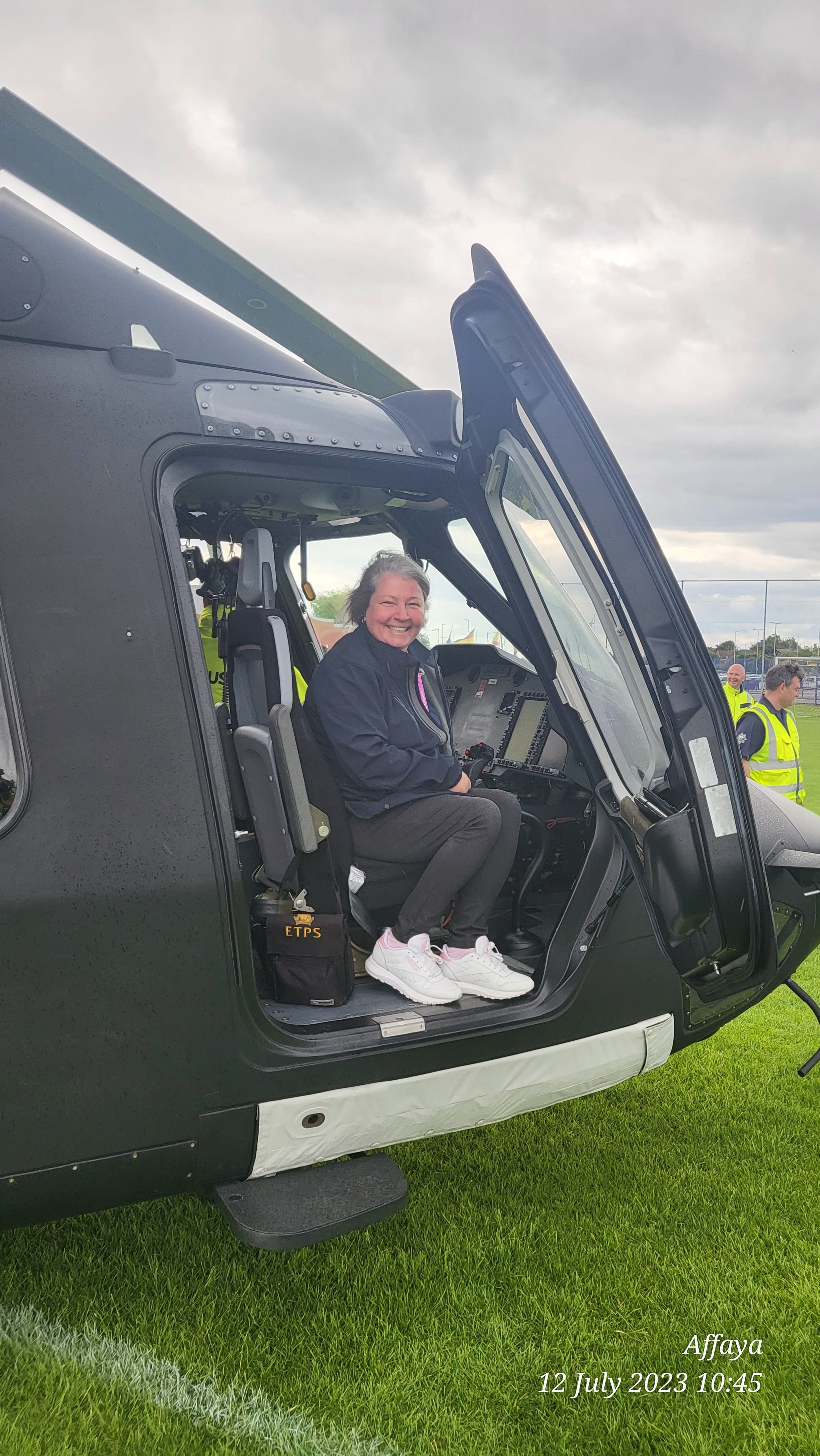 A smiling woman seated inside a helicopter cockpit on a grass field, with a cloudy sky overhead, and two men in yellow vests standing nearby. The photo is labeled with the location "Affaya" and the date "12 July 2023 10:45."