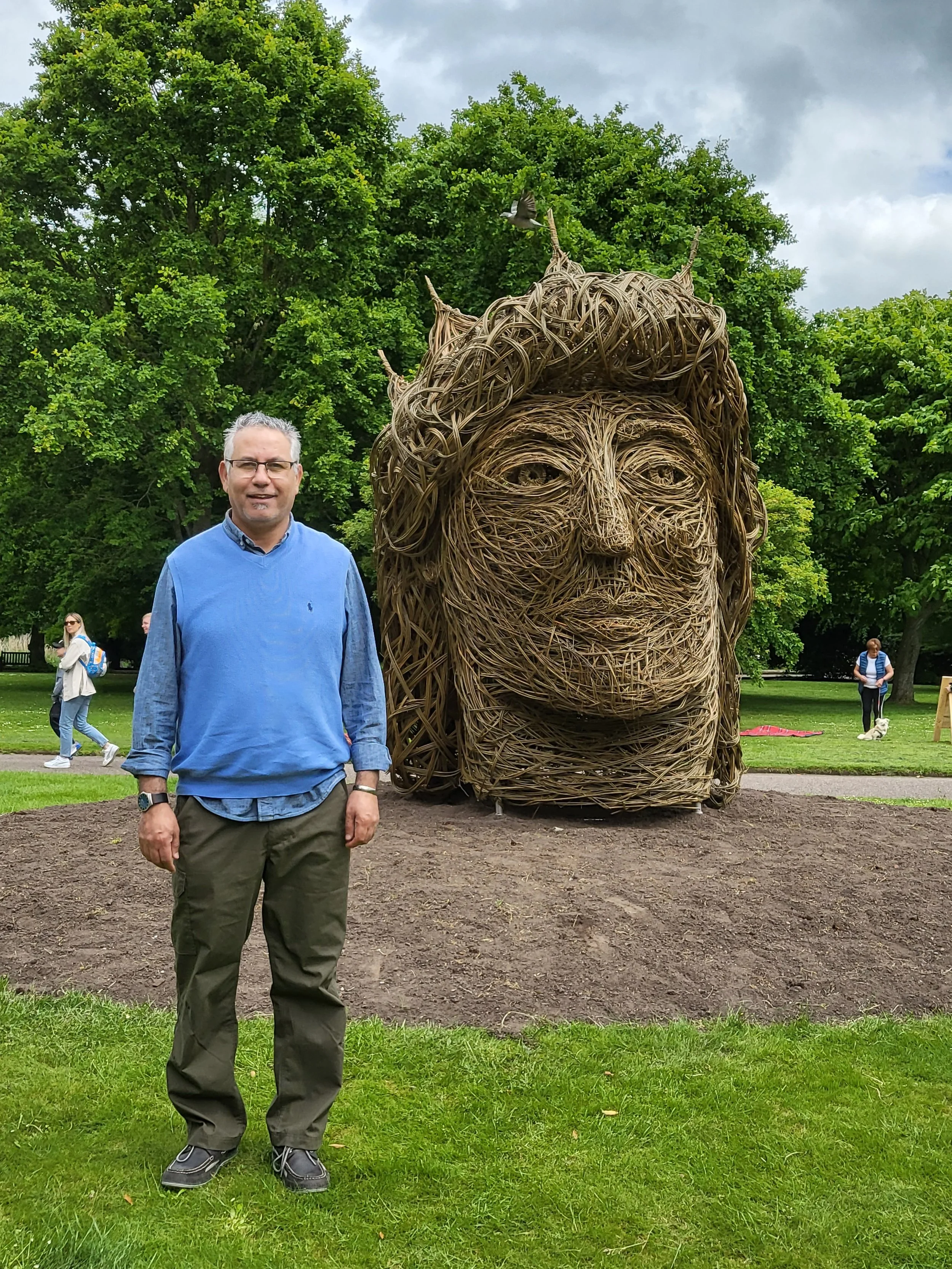 A man standing on a grassy area in front of a large outdoor sculpture of a woman's face made from intertwined wooden branches. There are trees and a few other people in the background.