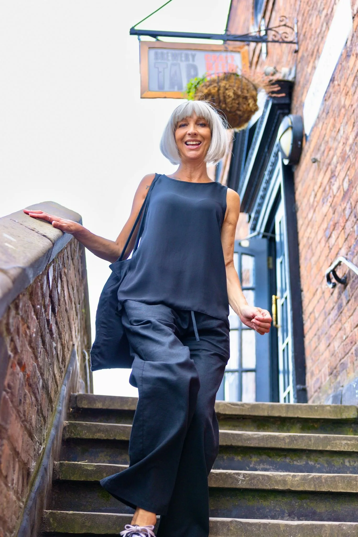 An older woman with gray hair, dressed in black, smiling and walking up outdoor stone stairs beside a brick building with a sign reading "Discovery Tap" and hanging plant.