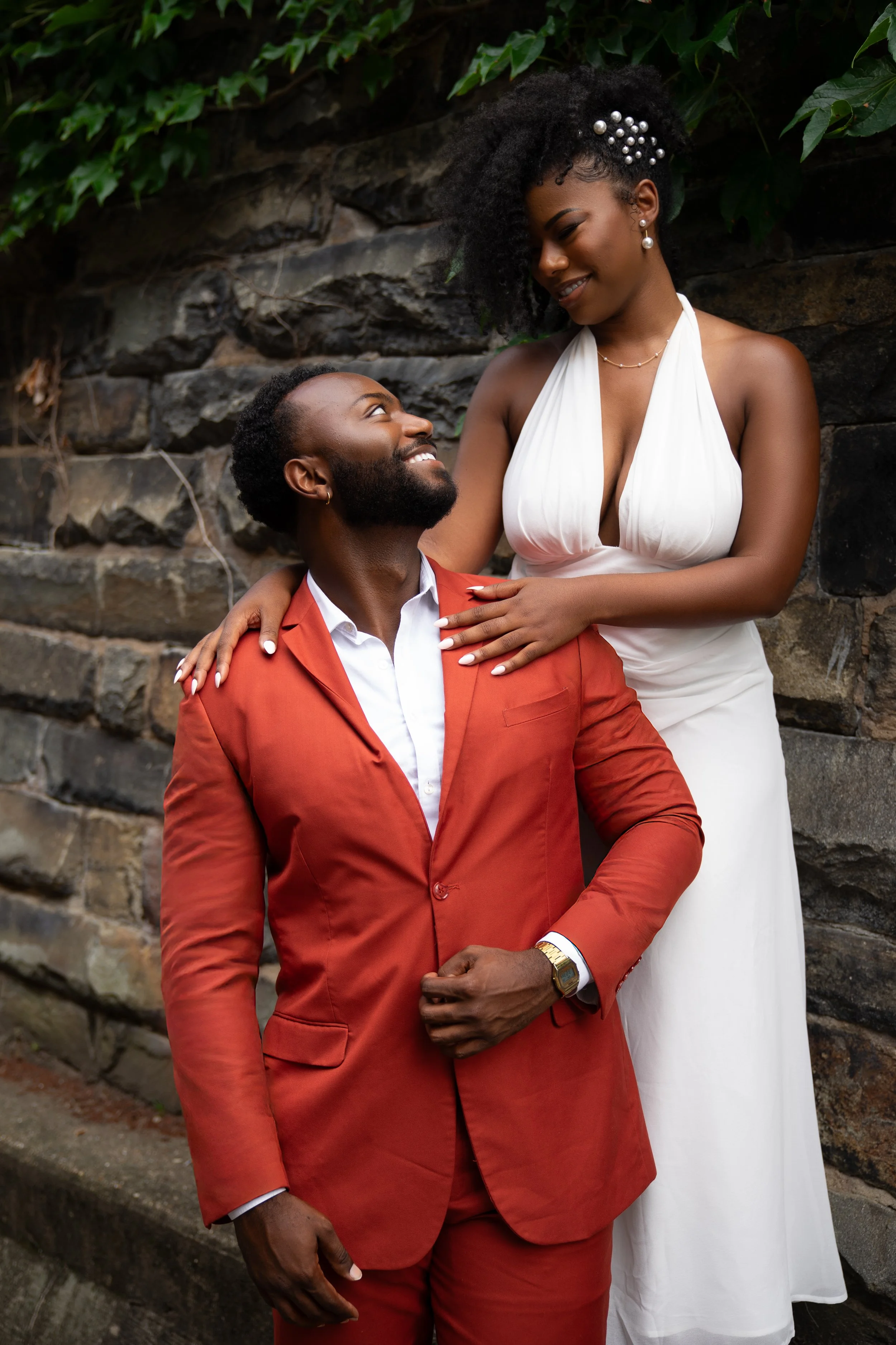 A couple dressed in formal attire, the woman in a white dress and the man in an orange suit, sharing a loving moment outdoors against a stone wall with green foliage.